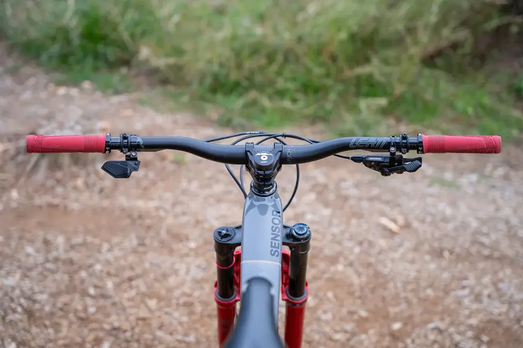 Vista aérea del manillar de una bicicleta de montaña, en la que destacan los puños ReaFlex 6.0 en un llamativo color rojo, las palancas de freno, y una palanca de cambio electrónica montada en un manillar Leatt Launch Control 6.0, con una potencia Gravity 6.0 que completa el conjunto; todo ello situado en un camino de tierra con una exuberante vegetación al fondo.