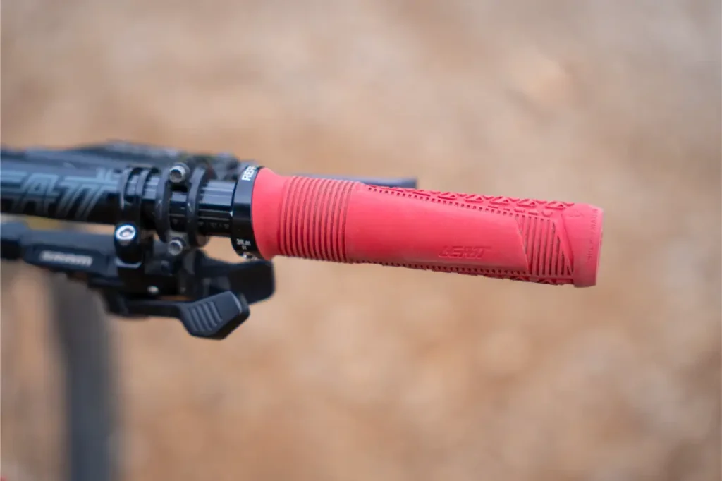 Primer plano del manillar de una bicicleta con un puño rojo de Leatt y la palanca de freno visible, en el que se aprecia el manillar Leatt Launch Control 6.0, sobre un fondo marrón difuminado.