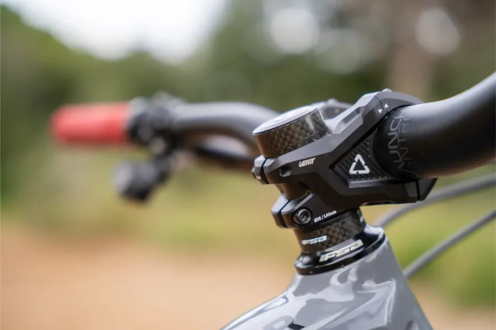 Primer plano del manillar y la potencia de una bicicleta de montaña, en el que destaca un puño ReaFlex 6.0 de un rojo intenso a la izquierda, todo ello montado en la robusta potencia Gravity 6.0, con un fondo natural desenfocado que aporta profundidad a la escena.
