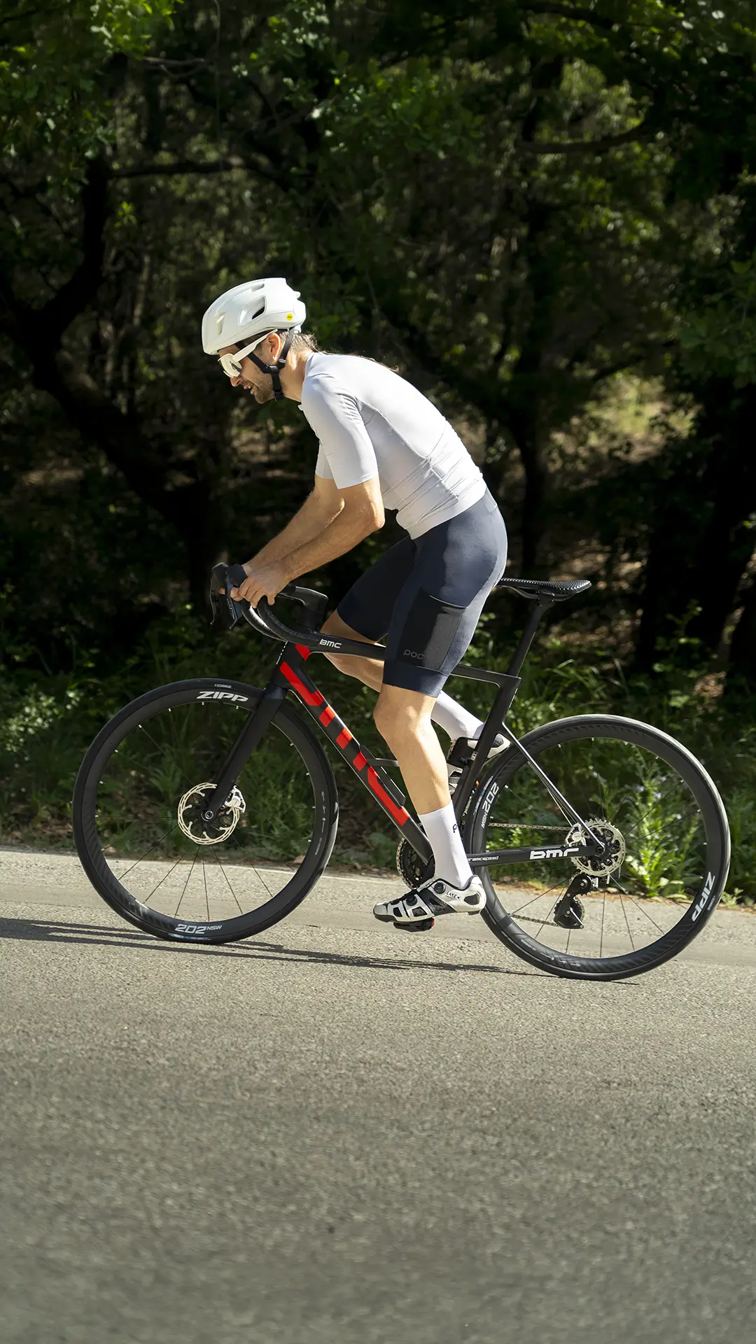 Un ciclista que lleva un casco blanco y un maillot de POC Cadence circula en una bicicleta de carretera negra y roja por una carretera asfaltada, disfrutando de la comodidad al pedalear, con árboles y vegetación al fondo.