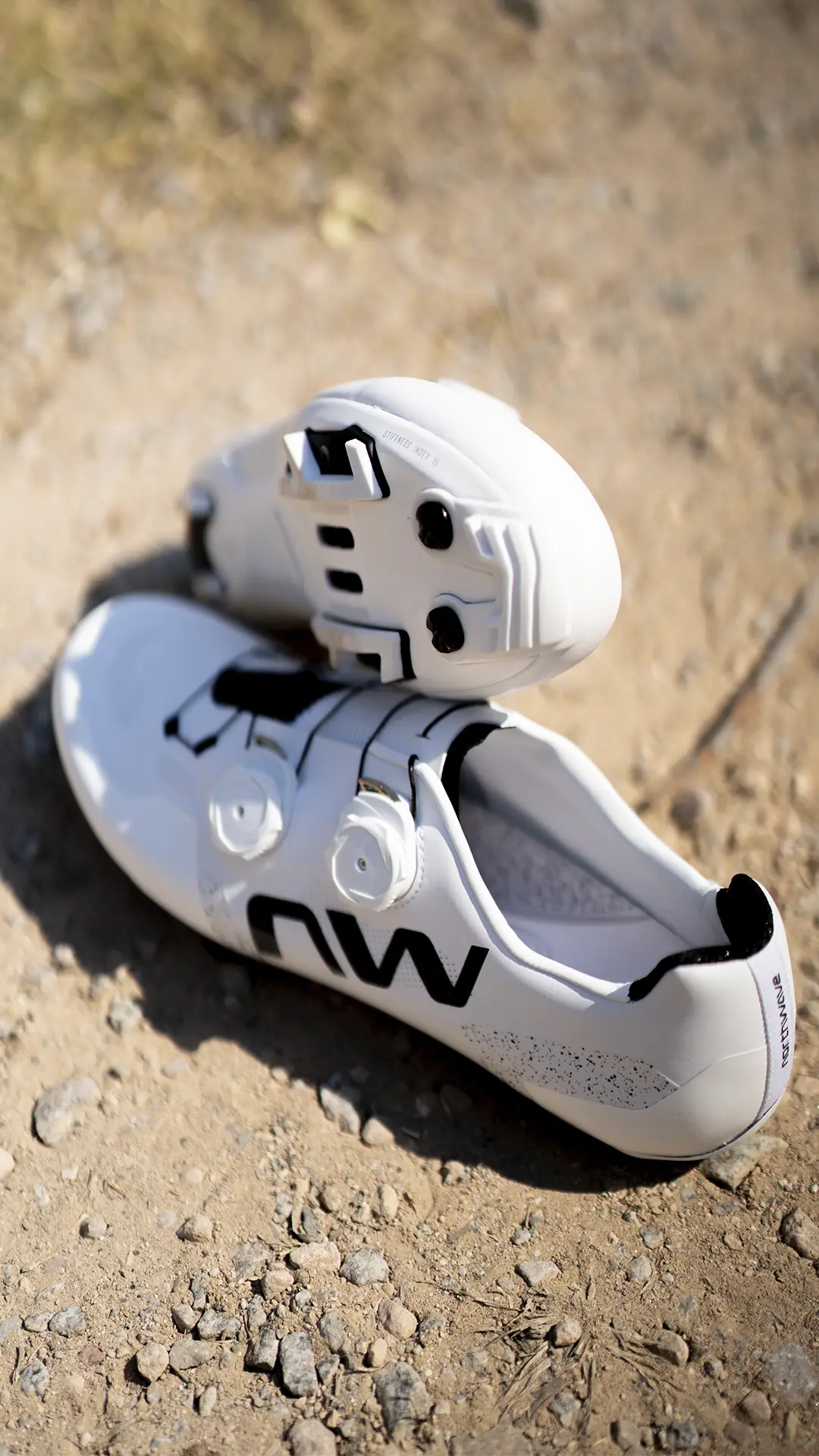 Un par de zapatillas de ciclismo Northwave Extreme X blancas con detalles en negro y cierres de dial, fotografiadas sobre una superficie de grava con una zapatilla apoyada de lado para mostrar la suela: una excelente opción para los ciclistas que buscan versatilidad y rendimiento en un modelo de gama alta.