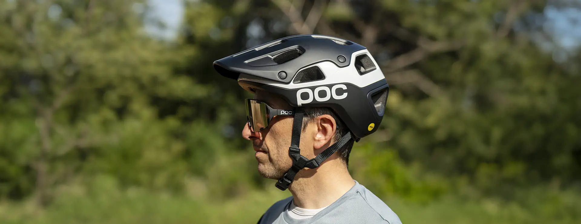 Un hombre que lleva un casco de ciclista POC Cularis Pure blanco y negro, con visera y gafas de sol, está de pie al aire libre, preparado para protegerse del sol, con un fondo verde difuminado.