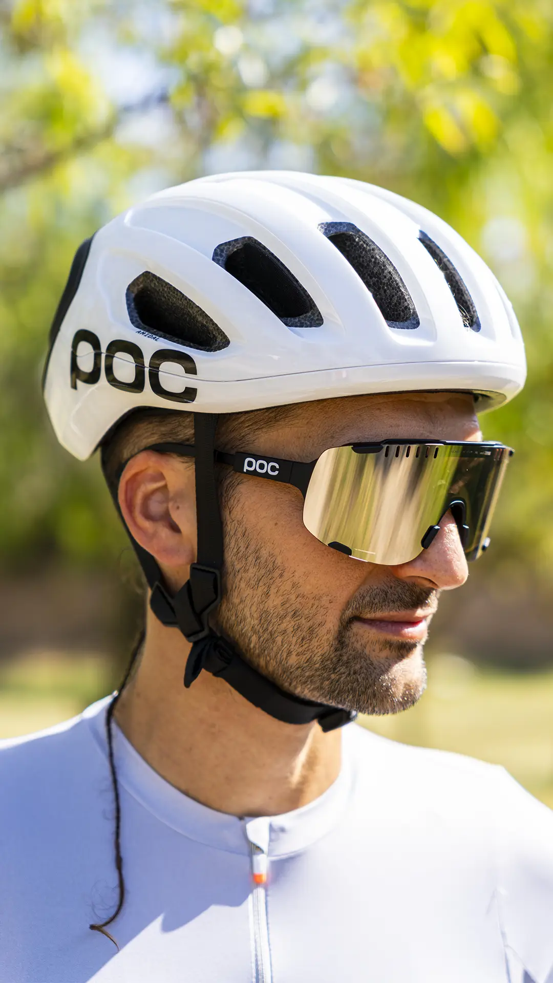 Hombre al aire libre con un casco de ciclismo polivalente blanco de la marca POC y unas gafas de sol grandes reflectantes, vestido con un maillot de ciclismo ligero adecuado para el ciclismo de carretera o gravel, con vegetación difuminada al fondo.