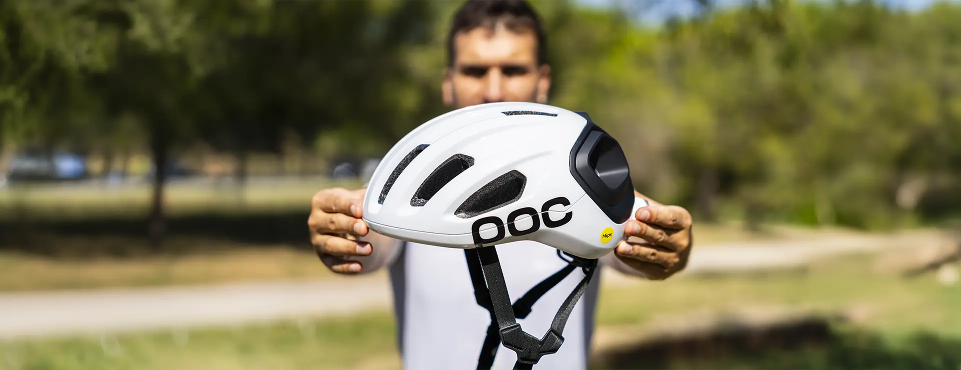 Una persona sostiene al aire libre un casco de ciclismo POC blanco con rejillas de ventilación negras y una correa ajustable, con árboles y césped al fondo; ideal como casco polivalente tanto para aventuras en grava como en carretera.