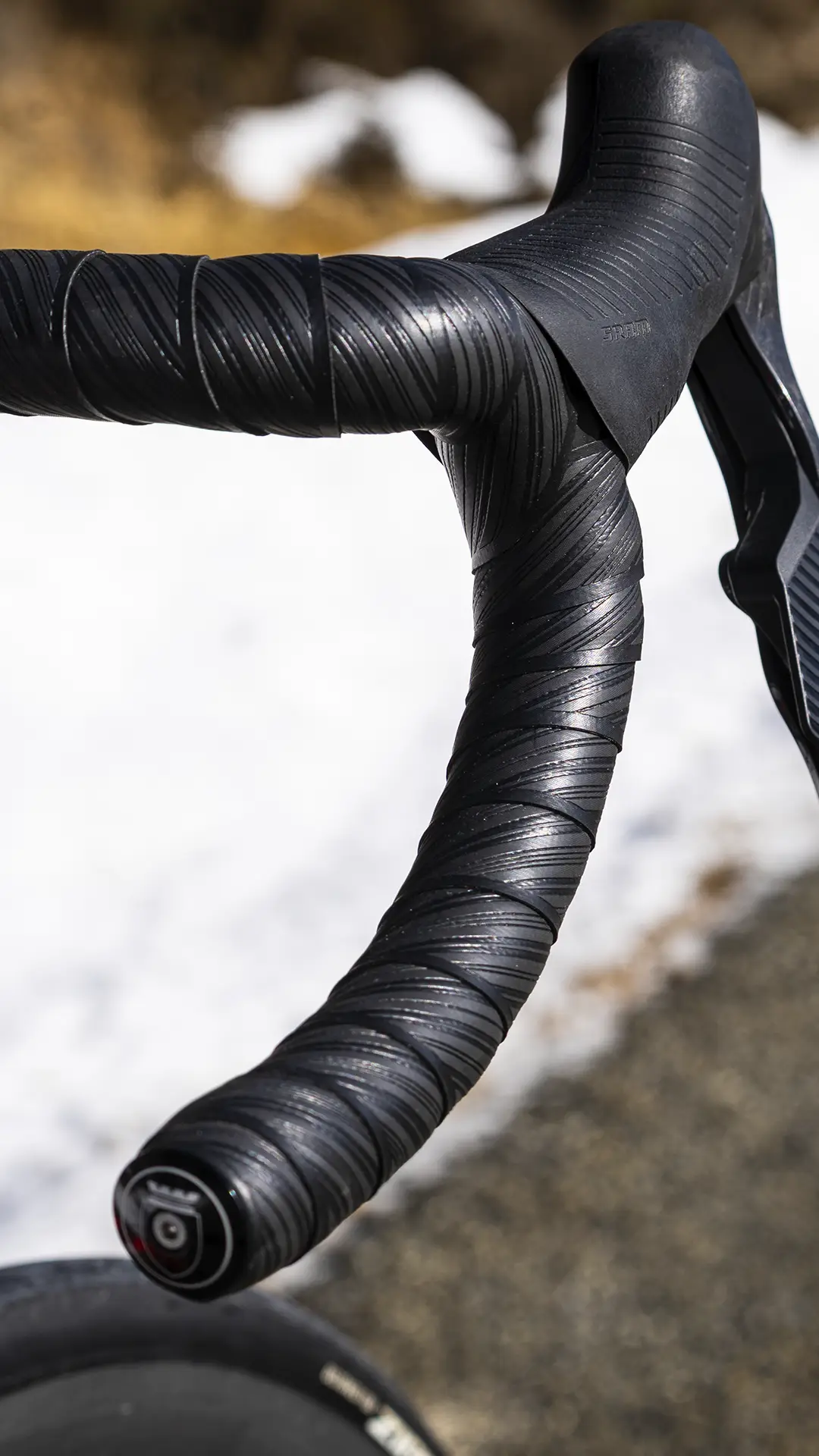 Primer plano de un manillar negro de bicicleta de carretera forrado con la cinta de manillar Silca Nastro Cuscino, una cinta texturizada diseñada para ofrecer mayor comodidad, fotografiado al aire libre junto a un suelo cubierto de nieve y una superficie de asfalto.