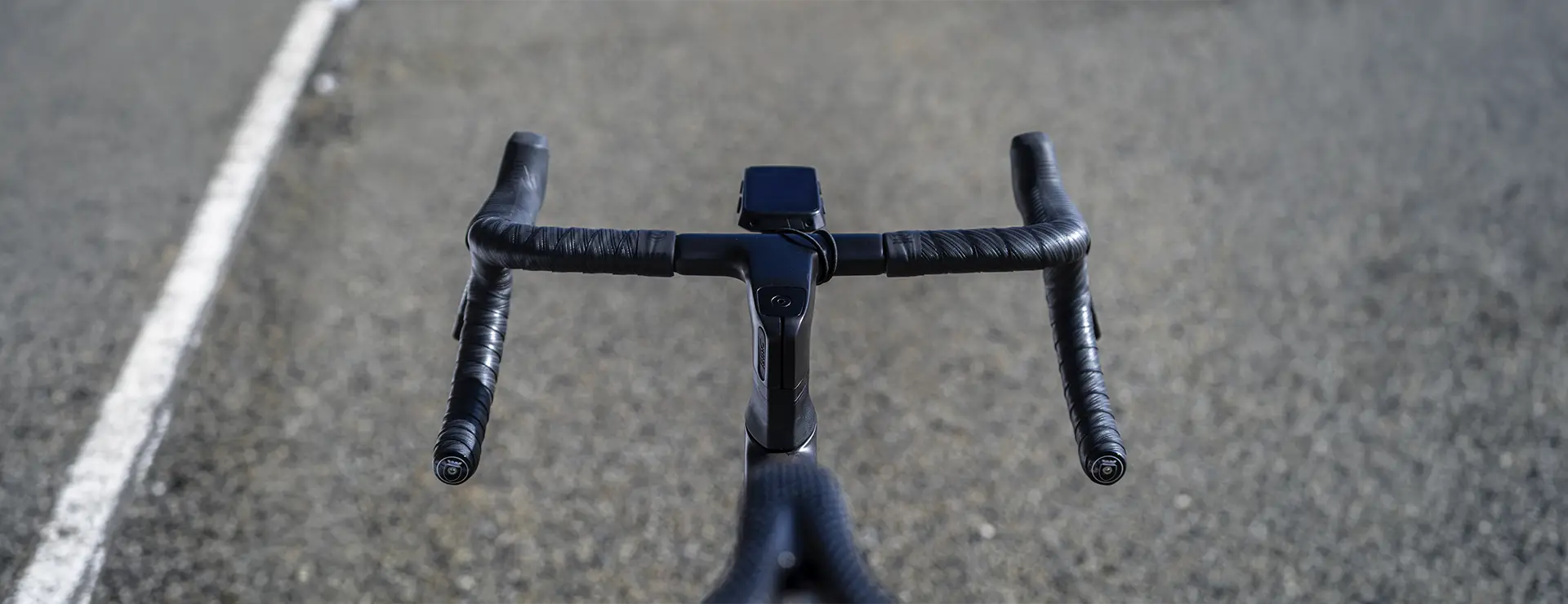 Primer plano del manillar de una bicicleta negra envuelto con cinta Silca Nastro Cuscino para mayor comodidad y tacto, con un ciclocomputador montado en el centro, en una carretera asfaltada y desierta.