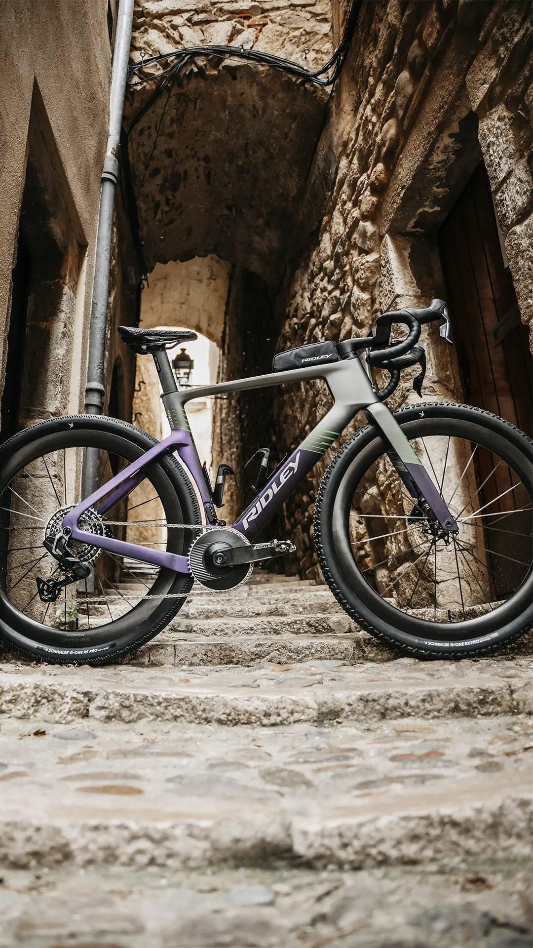 Una elegante bicicleta de carretera, la Ridley Kanzo Fast 2.0, con un llamativo cuadro morado y gris, está aparcada en una escalera de adoquines entre antiguos edificios de piedra. Su diseño aerodinámico destaca en medio del paisaje histórico, lista para deslizarse por cualquier ruta con estilo y velocidad.