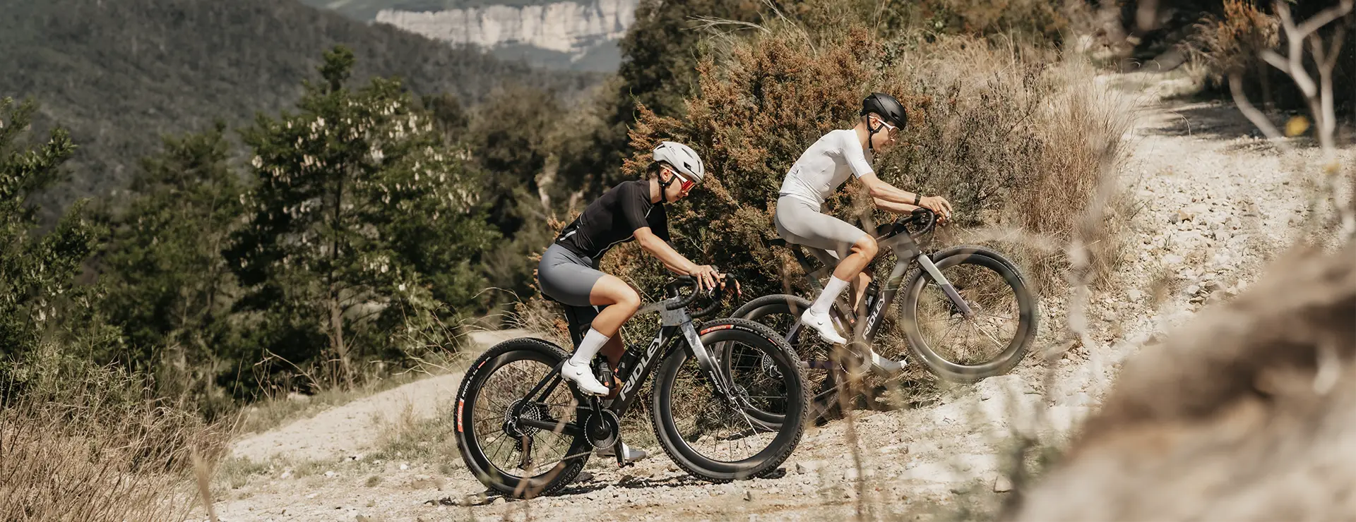 Dos ciclistas, con casco y ropa deportiva, suben en sus bicicletas de montaña Ridley Kanzo Fast 2.0 por un sendero de grava rodeado de árboles y colinas, afrontando con seguridad el terreno gracias a sus cuadros aerodinámicos y a los neumáticos de 58 mm, que les proporcionan mayor estabilidad.
