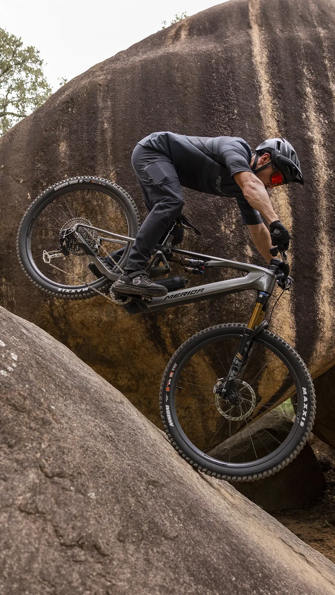 Un ciclista, vestido con ropa negra y casco, baja en bicicleta de montaña por una pendiente rocosa y empinada, manteniendo el equilibrio con la rueda delantera levantada.