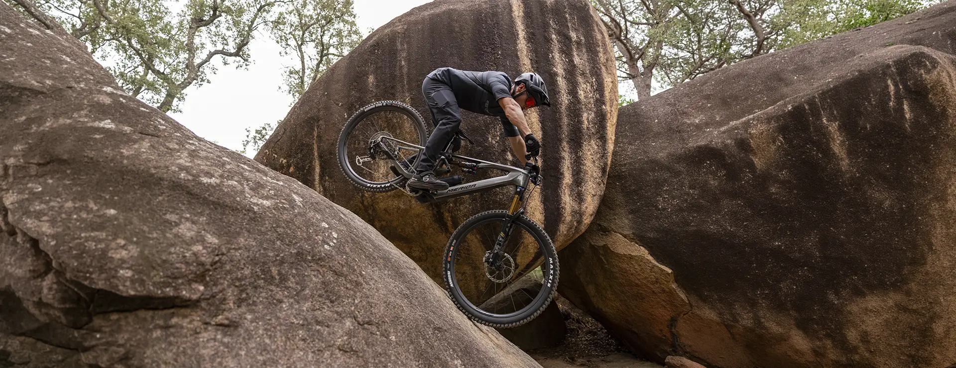 Un ciclista de montaña, con casco, sube por una empinada pared rocosa entre grandes cantos rodados en una zona boscosa.