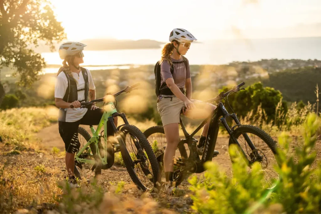 Dos personas con casco posan junto a sus bicicletas de montaña SCOTT Strike 2027 en un sendero de tierra, con vistas a un paisaje en el que se aprecian vegetación y agua al fondo durante la puesta de sol. Gracias al potente motor Bosch CX y a su excepcional comodidad, están listas para afrontar cualquier aventura.