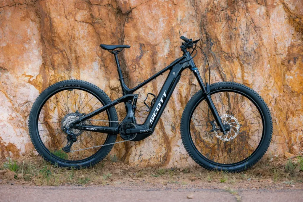 Una bicicleta de montaña eléctrica SCOTT Strike 2027 de color negro, con neumáticos anchos y un potente motor Bosch CX, está aparcada en posición vertical junto a una pared rocosa de color naranja y de aspecto rugoso, en un camino de tierra.