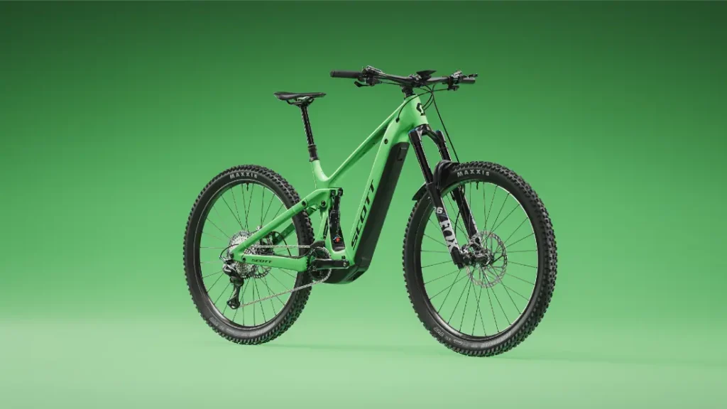 Una bicicleta de montaña verde con suspensión total, como la SCOTT Strike 2027 con motor Bosch CX, ofrece tanto comodidad como rendimiento gracias a sus neumáticos anchos y sus potentes frenos de disco, que resaltan maravillosamente sobre un fondo verde a juego.