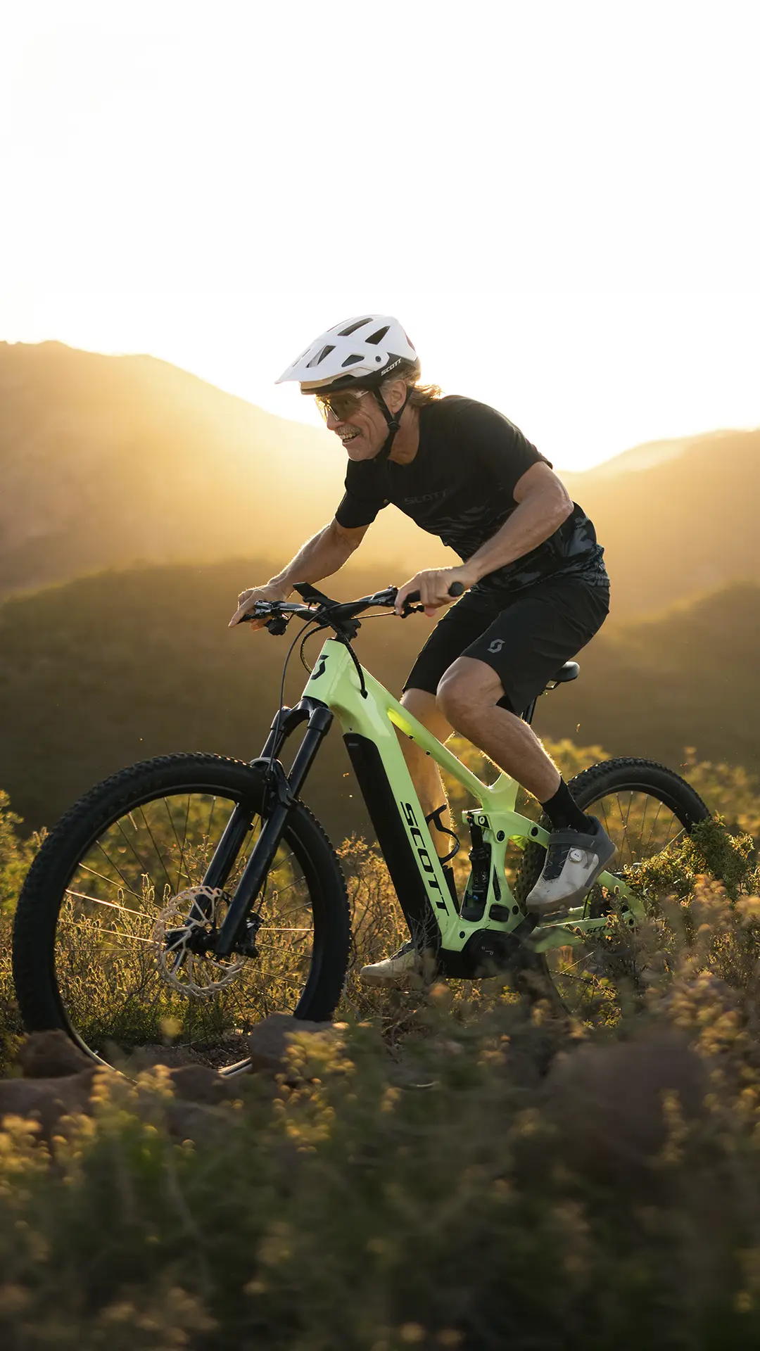 Un hombre con casco pedalea en una bicicleta de montaña SCOTT Strike 2027, equipada con un potente motor Bosch CX, por un sendero con colinas y la luz del sol al fondo, disfrutando tanto del rendimiento como de la comodidad durante su recorrido.