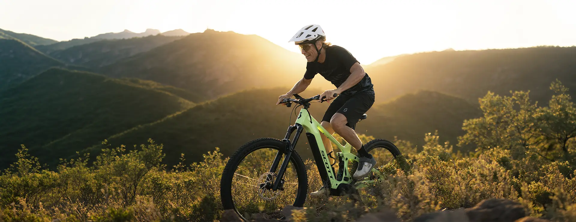 Una persona con casco monta en una bicicleta de montaña SCOTT Strike 2027, con una suspensión de 150 mm y un potente motor Bosch CX, subiendo por un sendero, con las montañas iluminadas por el sol al fondo.