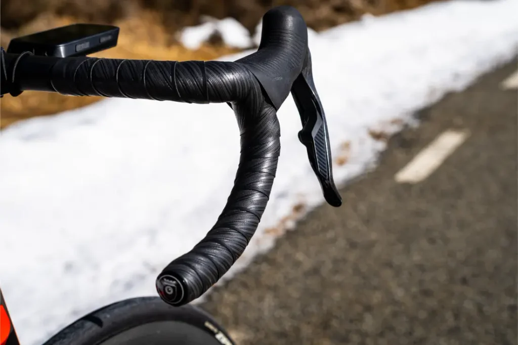 Primer plano del manillar negro de una bicicleta de carretera, envuelto con cinta Silca Nastro Cuscino, y de la palanca de freno, en una carretera asfaltada con nieve en los márgenes, lo que pone de relieve la excepcional comodidad y el tacto superior en cada salida.