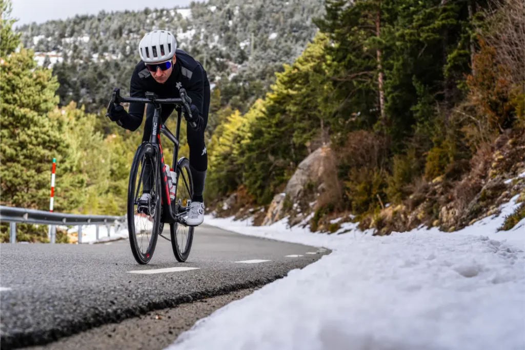 Un ciclista, con casco y gafas de sol, sube por una carretera de montaña, disfrutando de una comodidad excepcional y un tacto mejorado gracias a la cinta de manillar Silca Nastro Cuscino, con nieve a los lados de la carretera y árboles al fondo.