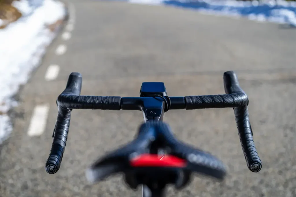 Primer plano del manillar de una bicicleta forrado con cinta Silca Nastro Cuscino y del sillín, con una carretera asfaltada al fondo. Se ve nieve a lo largo del borde de la carretera, lo que pone de relieve la necesidad de mayor comodidad y tacto durante los recorridos en días fríos.