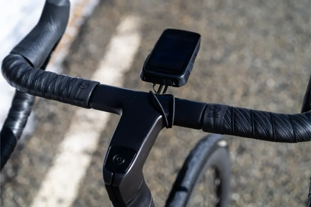 Primer plano del manillar de una bicicleta forrado con cinta Silca Nastro Cuscino negra, que ofrece un tacto y una comodidad excepcionales, con un ciclocomputador montado en una carretera al aire libre.