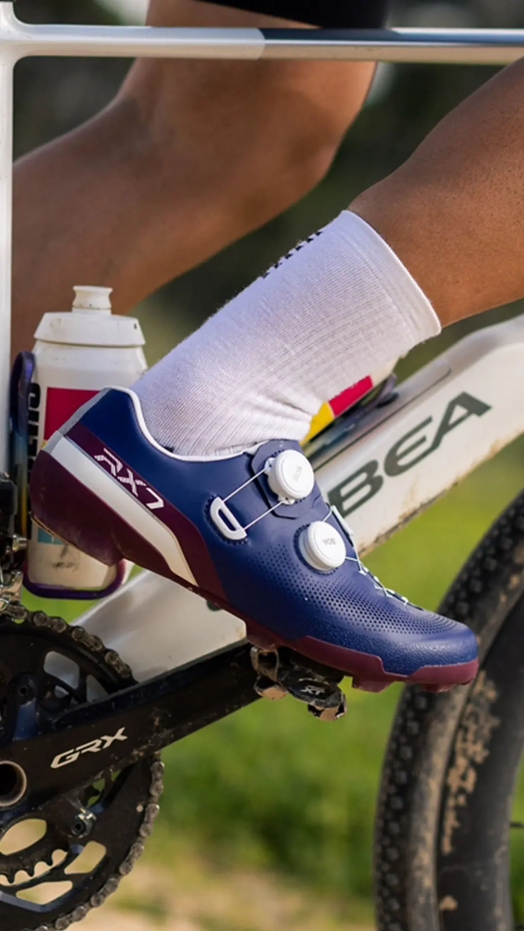 Primer plano del pie de un ciclista que lleva unas zapatillas azules Shimano RX710 en el pedal de una bicicleta, en las que se aprecia la tecnología de competición; también se ven un bidón y parte del cuadro blanco de una bicicleta ORBEA, ideal para cualquier aventura de gravel.