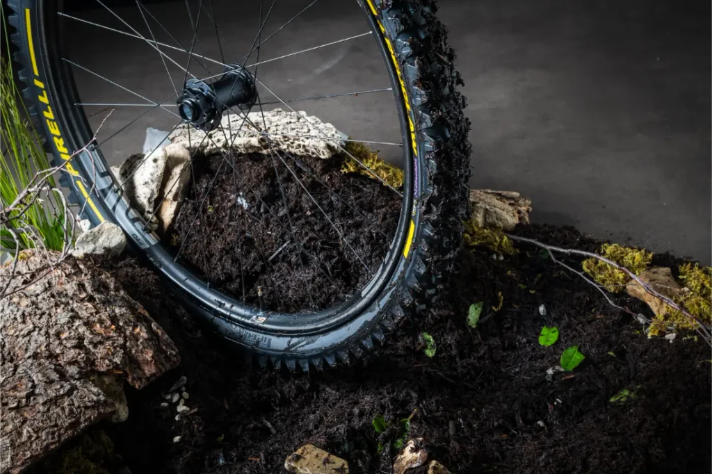 Primer plano de una rueda de bicicleta de montaña llena de barro, con el logotipo amarillo de Pirelli y la inscripción «Scorpion neumáticos MTB», apoyada sobre tierra, piedras y trozos de corteza en el interior de una habitación.