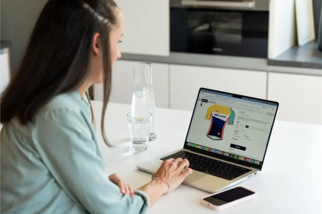 Una mujer está sentada a una mesa utilizando un ordenador portátil para comprar por Internet un maillot de ciclismo amarillo y azul. Sobre la mesa hay un vaso, una jarra de agua y un smartphone.