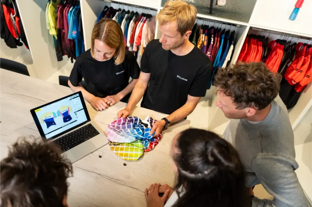 Cuatro personas observan un ordenador portátil en el que se muestran diseños gráficos a todo color y comentan muestras de tejidos en una tienda de ropa o un estudio de diseño, con diversas prendas colgadas en percheros al fondo.