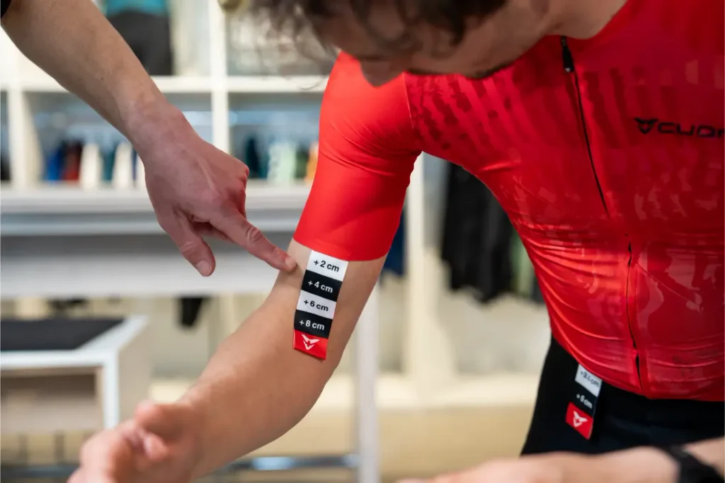 Una persona que lleva un maillot de ciclista rojo tiene una pegatina en el brazo con medidas en centímetros, mientras que otra persona señala la pegatina.