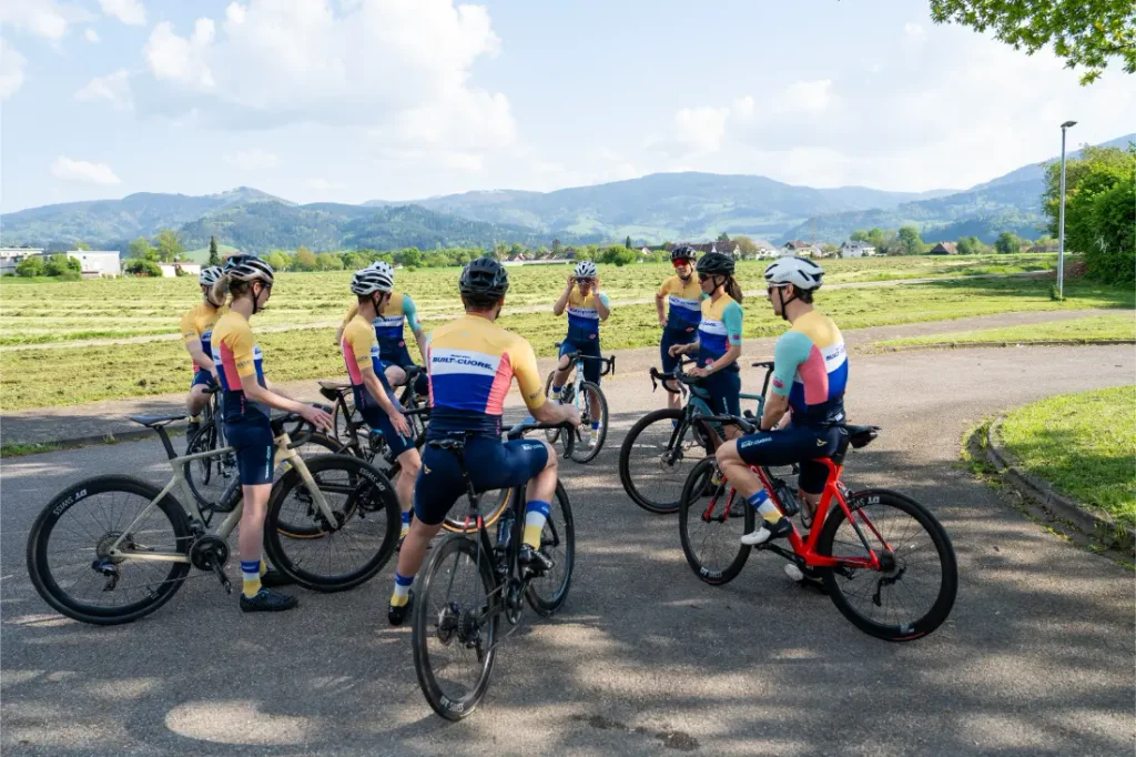 Ocho ciclistas, vestidos con uniformes a juego, se reúnen con sus bicicletas en un camino asfaltado, rodeados de campos y colinas bajo un cielo parcialmente nublado.