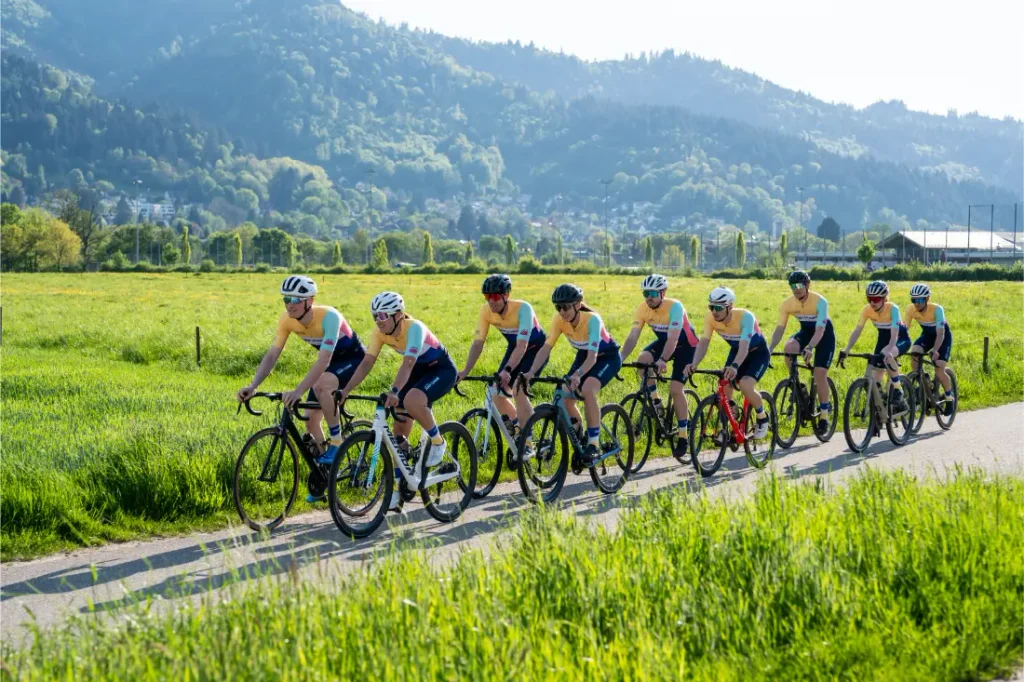 Nueve ciclistas, vestidos con equipación a juego, pedalean en fila por un camino asfaltado que atraviesa un prado verde, con las montañas al fondo.