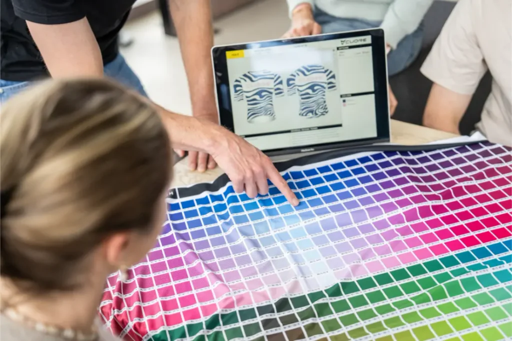 Un grupo examina una carta de muestras de telas de colores, mientras una persona señala un color y un ordenador muestra diseños de camisetas en la pantalla.