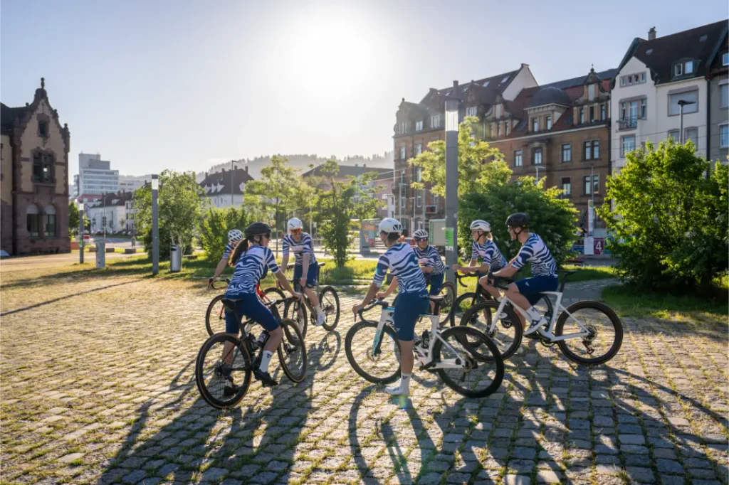 Un grupo de ciclistas con maillots a juego se reúne en círculo en una plaza empedrada, con la luz del sol y los edificios históricos como telón de fondo.