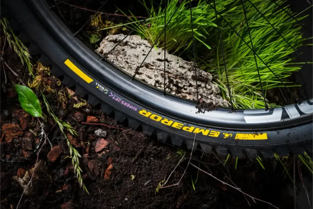Un primer plano de una rueda de bicicleta de montaña con la inscripción «EMP ERROR» sobre un terreno embarrado, con hierba verde y una roca cerca, en el que se muestran los neumáticos Scorpion MTB de Pirelli, que ofrecen una tracción superior.