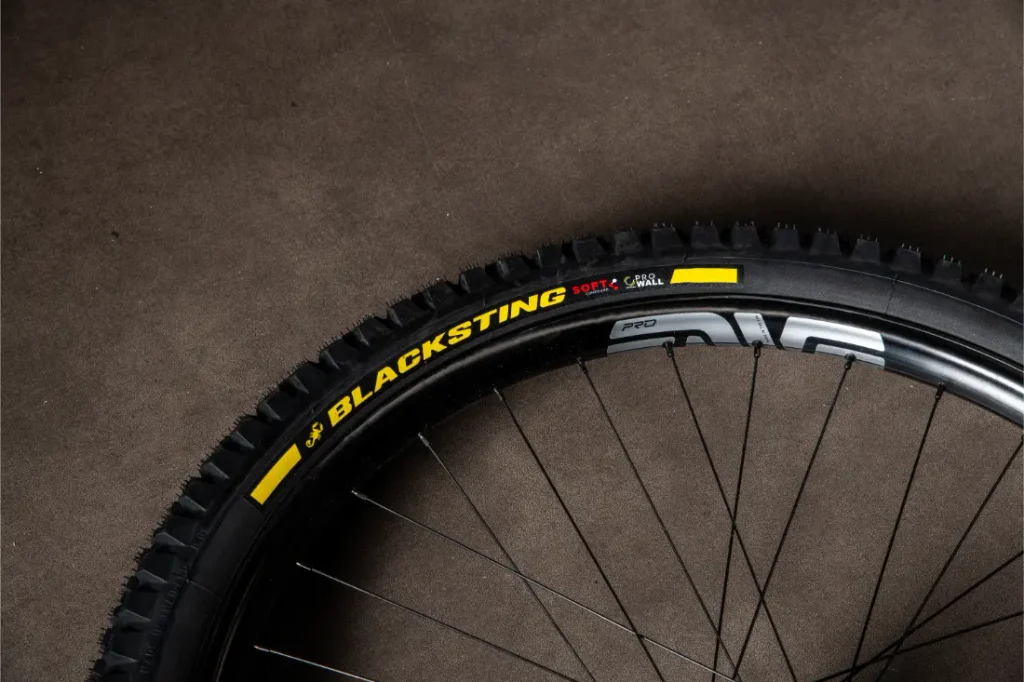 Un primer plano de una cubierta de bicicleta negra con la inscripción «BLACKSTING» en letras amarillas y rojas, montada en una rueda de radios, descansa sobre una superficie marrón lisa, lo que evoca el rendimiento resistente de los neumáticos de MTB de alta gama.