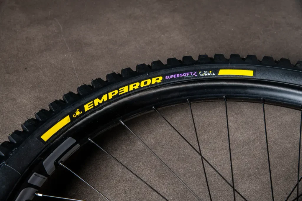 Primer plano de una cubierta de bicicleta negra con la etiqueta amarilla «EMPEROR» y los textos «SUPERSOFT» y «G-WALL» en el flanco, apoyada sobre una superficie oscura, que recuerda a los neumáticos Scorpion MTB de gama alta de Pirelli.
