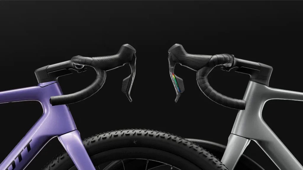 Dos manillares de bicicleta de carretera, uno morado y otro gris, aparecen colocados uno al lado del otro sobre un fondo negro, con las ruedas delanteras y las palancas de freno a la vista. Esta composición evoca el espíritu polivalente de la SCOTT Addict Gravel 2027, que ofrece un agarre cómodo para largas rutas por terrenos variados.