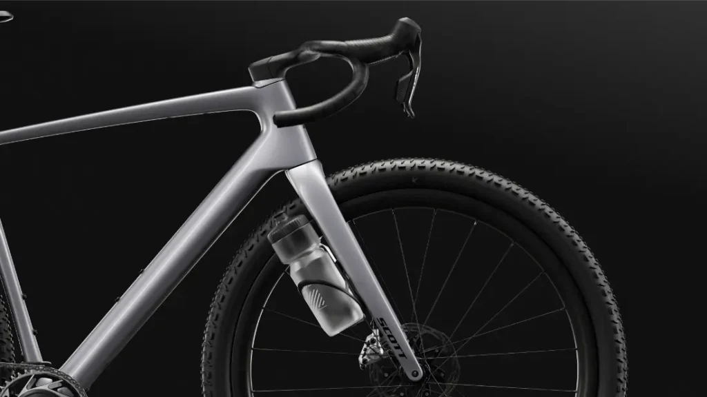 Primer plano de una moderna SCOTT Addict Gravel 2027 de color gris, centrado en la parte delantera, en el que se aprecian el manillar curvo, el bidón y la rueda con textura sobre un fondo negro: una cómoda bicicleta de gravel ideal para cualquier aventura.