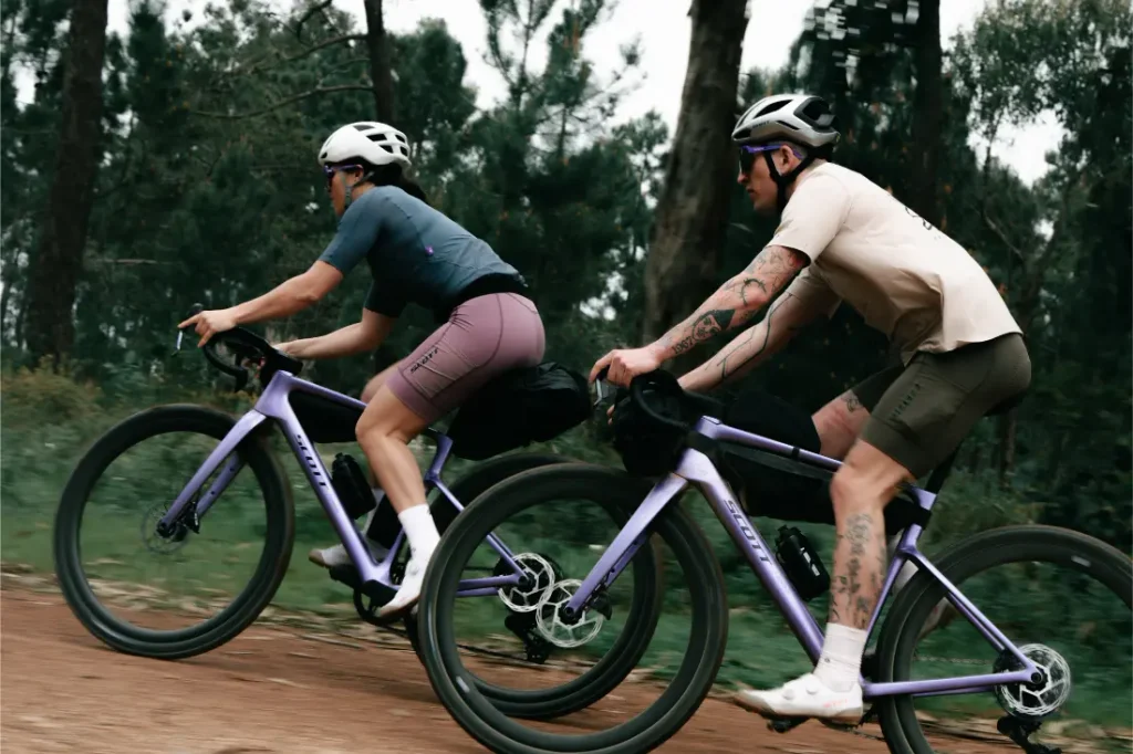 Dos ciclistas, equipados con casco y ropa deportiva, pedalean en unas bicicletas SCOTT Addict Gravel 2027 de color morado por un camino de tierra que atraviesa una zona boscosa, disfrutando de una experiencia ciclista cómoda y versátil.