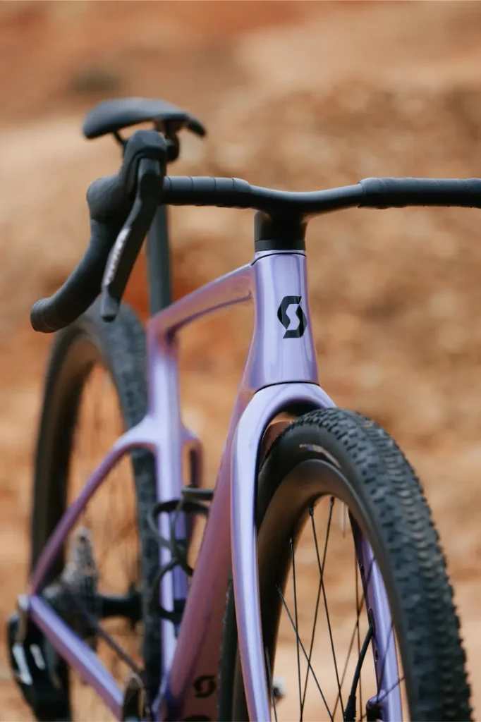 Primer plano de una bicicleta SCOTT Addict Gravel 2027 de color morado, con manillar de carretera y neumáticos de tacos, sobre un fondo difuminado en tonos tierra. Esta bicicleta de gravel polivalente ofrece una experiencia de conducción cómoda en terrenos variados.