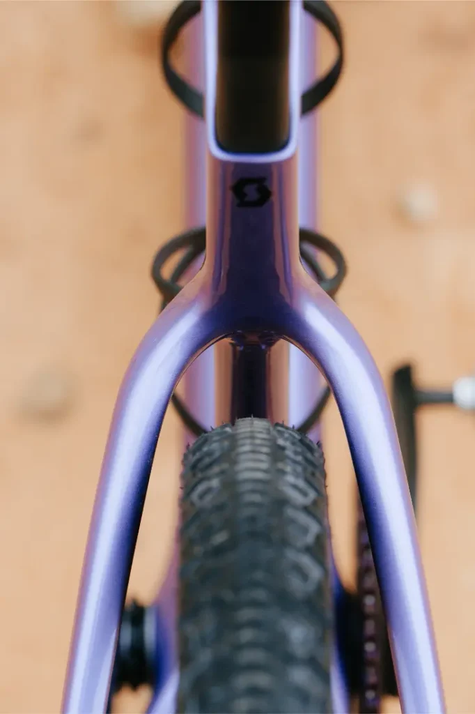 Primer plano desde arriba de un cuadro morado de bicicleta de gravel, con especial atención a la rueda trasera y a los tirantes del sillín, y con un suelo de tierra difuminado al fondo: un aspecto emblemático para una bicicleta polivalente como la SCOTT Addict Gravel 2027.