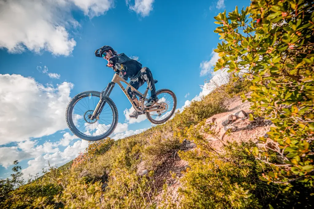 Una persona en bicicleta de montaña se lanza desde un sendero de tierra en un paisaje montañoso, bajo un cielo azul salpicado de nubes blancas.