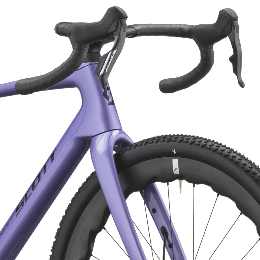 Primer plano de una bicicleta SCOTT Addict Gravel 2027 de color morado en el que se aprecian el manillar curvo, las palancas de freno delanteras y la rueda delantera con banda de rodadura de tacos: características perfectas para una «gravel cómoda» y una bicicleta polivalente.