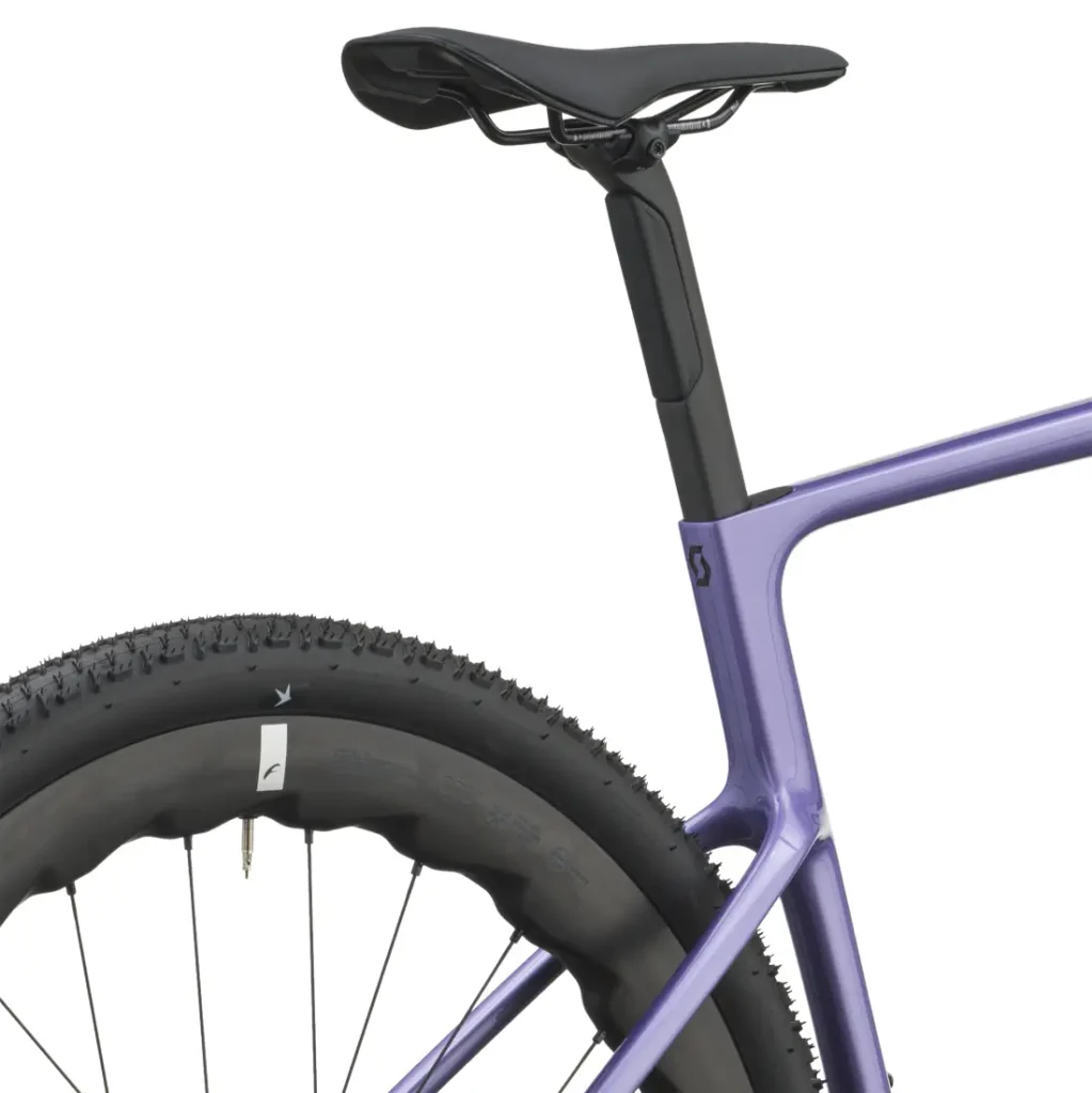 Primer plano de un sillín negro, una tija de sillín y una rueda trasera, en el que se aprecia el cuadro morado de la bicicleta SCOTT Addict Gravel 2027 y la banda de rodadura de los neumáticos con tacos: detalles perfectos para quienes buscan una bicicleta cómoda y polivalente.