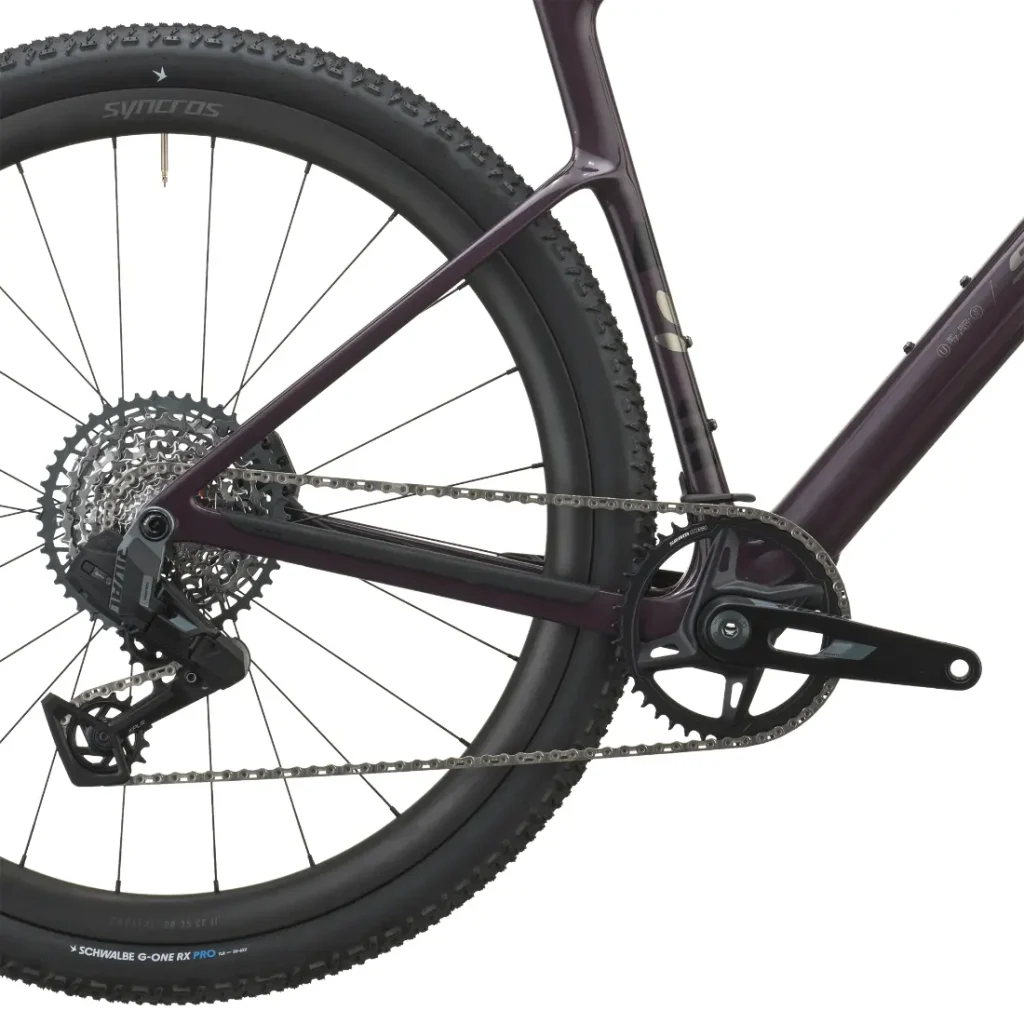 Primer plano de la rueda trasera, la cadena, los cambios y el juego de bielas de una SCOTT Addict Gravel 2027. El cuadro es de color morado oscuro y el neumático lleva la inscripción «Schwalbe G-One R». Esta bicicleta de gravel, polivalente y cómoda, ofrece un equilibrio perfecto entre rendimiento y versatilidad gracias a sus componentes de alta calidad.