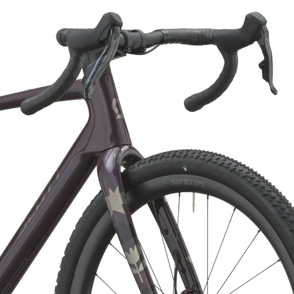 Primer plano de la rueda delantera, la horquilla y el manillar curvo de una bicicleta SCOTT Addict Gravel 2027 de color oscuro, con neumáticos texturizados y la marca «SYNCROS» en la llanta, lo que pone de manifiesto su diseño cómodo y versátil.