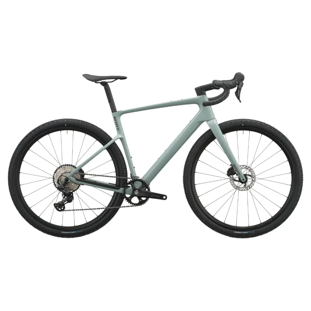 Se muestra de perfil, sobre un fondo blanco, una bicicleta de gravel SCOTT Addict Gravel 2027 de color verde mate, con manillar curvo, sillín negro y neumáticos grandes y con tacos, lo que resalta su diseño polivalente.