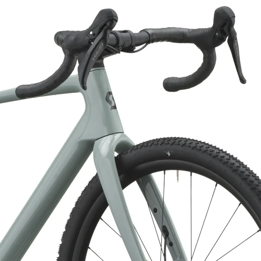 Primer plano de una bicicleta SCOTT Addict Gravel 2027 de color gris claro en el que se aprecian la rueda delantera, la cubierta con banda de rodadura de tacos, ideal para recorridos por grava, la horquilla, el manillar y las manetas de freno. Esta bicicleta polivalente está diseñada para rendir al máximo en una gran variedad de terrenos.