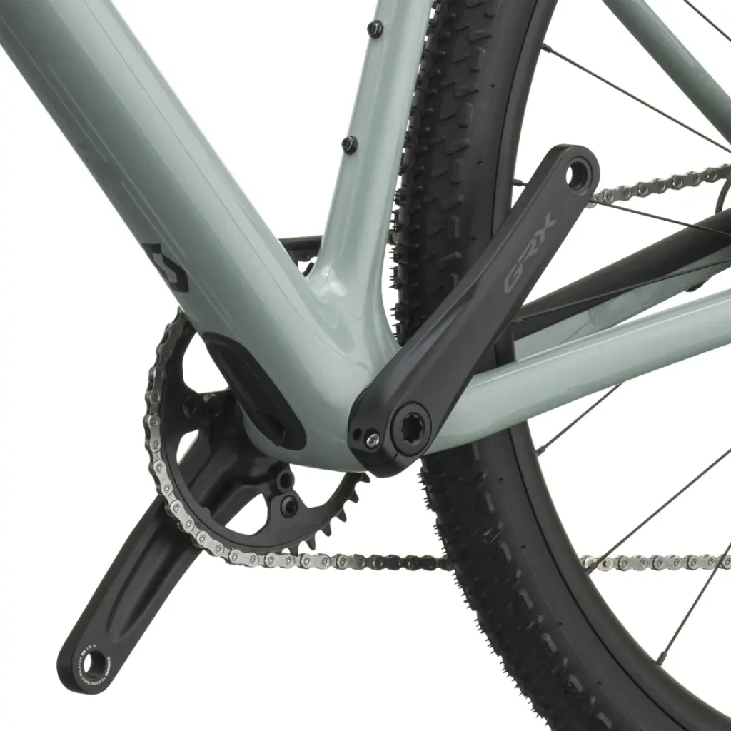 Primer plano de un juego de bielas, un plato y una rueda trasera de bicicleta, en el que destaca la biela GRX montada en un cuadro de bicicleta SCOTT Addict Gravel 2027 de color gris claro, con la banda de rodadura de los neumáticos de tacos bien visible. Esta bicicleta polivalente y cómoda está diseñada para ofrecer versatilidad y comodidad en cada salida.