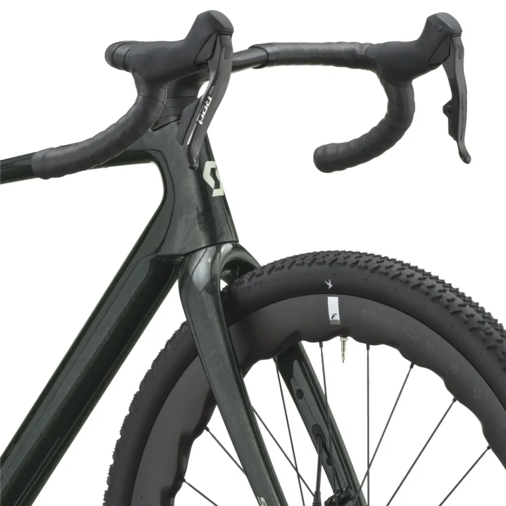 Primer plano de la rueda delantera, la horquilla y el manillar curvo de una cómoda bicicleta gravel, con cinta de manillar negra. La rueda presenta una banda de rodadura con tacos, y el cuadro lleva el logotipo «SCOTT Addict Gravel 2027».