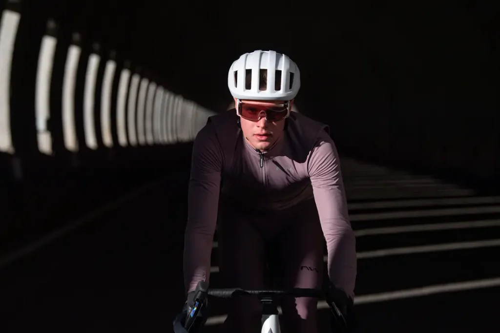 Un ciclista que lleva un casco blanco y gafas de sol atraviesa un túnel con patrones de luz a rayas en las paredes y el suelo.
